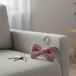 May include: A crocheted bow-tie keychain in a dusty rose color. The bow is attached to a silver-colored key ring with a short chain. A set of keys rests on the gray upholstered surface next to the keychain.