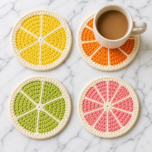 May include: Four handmade crochet coasters in the shape of citrus fruit slices. The coasters are yellow, orange, green, and pink, with white accents and a cream-colored border. One coaster holds a white mug with a brown beverage.