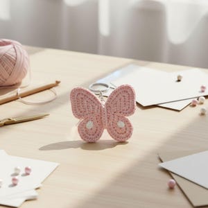 May include: A pink crocheted butterfly keychain with white accents. The handmade butterfly has a silver keyring attached. The keychain is on a light wooden surface with craft supplies, including yarn and scissors.