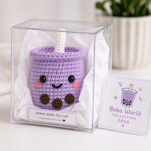 Puede incluir: Una taza de té boba morada tejida a mano, con una cara sonriente, rubor y perlas de boba marrones, presentada en una caja transparente. La etiqueta dice "Kawaii Boba Tea Cup" y "Handmade with Love". Una tarjeta con un gráfico de té boba dice "Boba World Collection 2026".