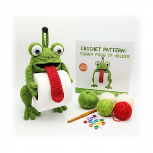 May include: A green crocheted frog toilet paper holder with a red tongue holding a roll of white toilet paper. The frog has large white eyes and a black antenna. A crochet pattern book and yarn are also in the image.
