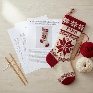 Può includere: Una calza di Natale lavorata a maglia con un motivo a fiocco di neve rosso e crema, con il nome "FINN" in alto. L'immagine include ferri da maglia, un uncinetto, filato e un modello intitolato "Nordic Snowflake Christmas Stocking".