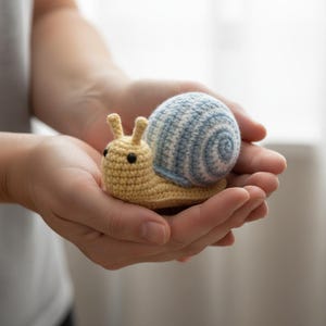 Puede incluir: Un juguete de caracol tejido a crochet. El cuerpo es amarillo claro, la concha a rayas azules y blancas. El caracol tiene ojos negros y dos pequeñas antenas. El juguete se sostiene en manos abiertas. Producto artesanal.