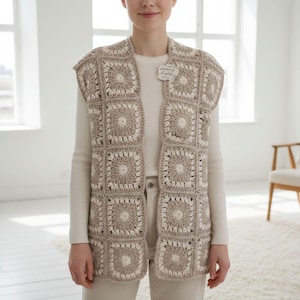 Puede incluir: Un chaleco de crochet beige y crema con un patrón cuadrado. El chaleco sin mangas es abierto por delante. Una pequeña etiqueta está adherida al chaleco. El chaleco se lleva sobre una camisa crema de manga larga.