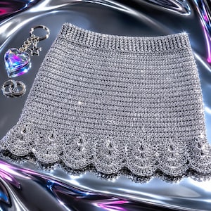 Op de afbeelding: Een metallic zilveren gehaakte rok met een gestructureerd breipatroon en een geschulpte zoom. Naast de rok liggen twee zilveren oorringen en een sleutelhanger met een hartvormige iriserende bedel en een kleine zilveren ster.
