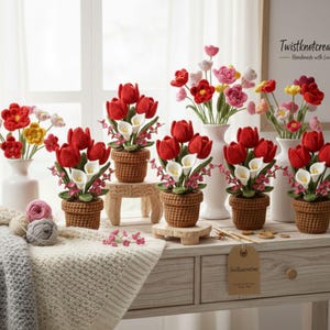 May include: Handmade crochet flower arrangements featuring red tulips and white calla lilies in brown woven pots. Other vases display colourful crochet flowers. The scene is set on a wooden table with yarn and a blanket.