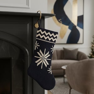 May include: Navy blue Christmas stocking with a white snowflake design and a zig-zag pattern at the top. The stocking is hanging from a gold-colored hook on a black fireplace mantel. The stocking is made of a knitted material.