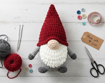 May include: A handmade crochet gnome with a red hat, white beard, and gray body. The gnome is surrounded by craft supplies, including yarn, buttons, and a tag that says "Made with Love".