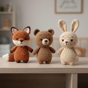 Puede incluir: Tres juguetes de animales de ganchillo: un zorro, un oso y un conejo. El zorro es naranja con cara y cola blancas y marrones. El oso es marrón y el conejo es beige. Los tres están sobre una superficie marrón claro.