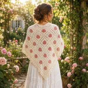 May include: A cream-colored, crocheted shawl with a geometric pattern of pink floral accents. The shawl is draped over the shoulders, with a scalloped edge. The background features a garden with pink roses and a white dress.