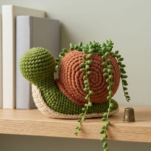 Puede incluir: Una maceta de caracol de ganchillo hecha a mano en verde, beige y marrón, con una planta suculenta colgante. La concha del caracol tiene un diseño en espiral. La maceta está sobre una estantería de madera.