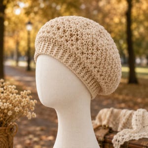 May include: A beige crochet beret displayed on a mannequin head. The beret has a textured, star-like pattern and a ribbed brim. The background is a blurred outdoor scene with fall foliage.