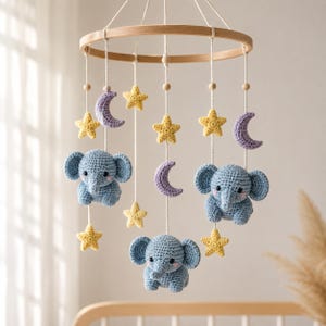 May include: A hanging mobile featuring three crocheted blue elephants, several yellow crocheted stars, and three purple crocheted crescent moons suspended from a circular wooden frame by white cords with small wooden beads.