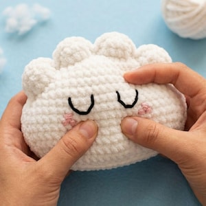 Peut inclure: Un jouet en peluche blanc en forme de nuage crocheté, avec des yeux cousus noirs et des joues roses. Le jouet doux et texturé est tenu par deux mains sur un fond bleu clair. Une pelote de laine blanche et un petit nuage sont également visibles.