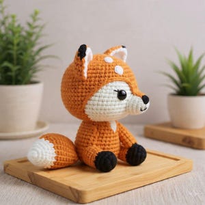 May include: A handmade crocheted fox toy in orange, white, and black. The fox features a white face and chest, an orange body, and a white-tipped tail. It is displayed on a wooden tray, highlighting the intricate crochet work.