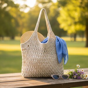 May include: A beige crochet tote bag with long handles, a straw hat, and a blue scarf. The bag is on a wooden picnic table with sunglasses and a small bouquet of flowers. The background is a blurred green park.