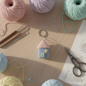May include: A handmade blue and pink crochet house keychain with a yellow button and a small green door. The keychain is surrounded by balls of yarn in various pastel colors, crochet hooks, and a pair of scissors.