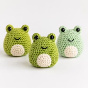 May include: Three handmade crochet frog toys in varying shades of green. Each frog has black eyes, a smiling mouth, and a cream-colored belly. The frogs are displayed against a white background.