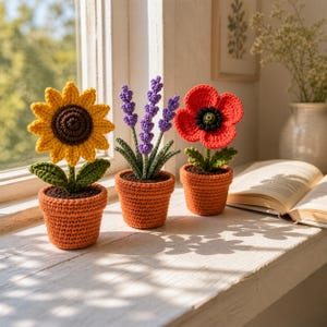 May include: Three crocheted flower plants in terracotta pots sit on a windowsill. A yellow sunflower, purple lavender, and red poppy are displayed. The flowers have green leaves and brown centers. The pots are orange and filled with brown soil.