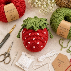 May include: A handmade crochet strawberry pouch with a green leaf top and white seed details. The pouch is surrounded by red and green yarn, a crochet hook, scissors, embroidery thread, and a needle. The yarn labels read "DK COTTON 100% Cotton".