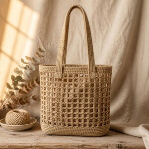 May include: A beige crochet tote bag featuring a structured rectangular base and an open grid pattern throughout the body. The bag includes two matching crochet shoulder straps and a solid beige fabric lining visible through the grid design.