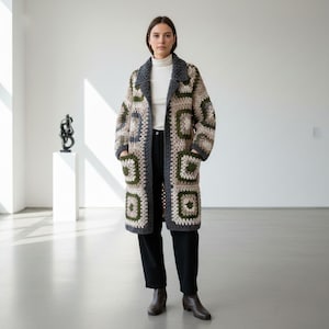 May include: A long, open crochet coat in a patchwork design. The coat features squares of beige, olive green, and grey yarn. It has a shawl collar and pockets. The person is wearing a white roll neck and black trousers.