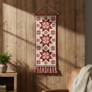 May include: A rectangular wall hanging with a cream background and red starburst patterns. The top has a wooden rod and the bottom has red and cream tassels. The wall hanging is suspended by a cream-colored string.
