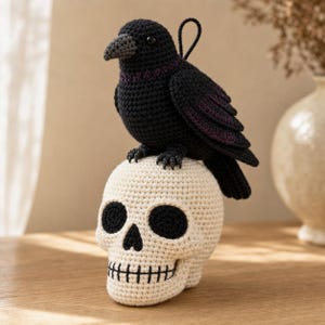 Puede incluir: Un cuervo de ganchillo hecho a mano con hilo negro y sutiles acentos morados posado sobre una calavera de ganchillo de color crema. El cuervo tiene un pequeño lazo en la espalda para colgarlo, mientras que la calavera tiene detalles en hilo negro.