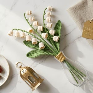 May include: A handcrafted bouquet of crochet lily of the valley flowers in a clear glass vase. The flowers are cream-colored with green stems and leaves. A gold-colored lantern and a plate are also visible.