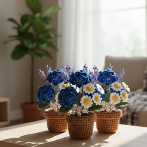 May include: Three crochet flower arrangements in small brown pots. The flowers include blue hydrangeas, white daisies with yellow centers, and purple lavender. The pots are sitting on a wooden table in front of a window.