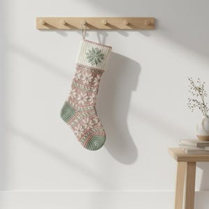 Op de afbeelding: Een kerstsok met een sneeuwvlokkenpatroon, in roze, groen en crème, hangt aan een houten kapstok. De sok heeft een gebreid ontwerp met een groene sneeuwvlok op de manchet. De teen en hiel zijn groen.