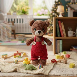 Puede incluir: Un oso de peluche de ganchillo marrón hecho a mano con un peto rojo con botones blancos. El oso tiene un hocico blanco y ojos verdes. Varios juguetes de madera, incluyendo un tren, un pato amarillo y un dinosaurio verde, están colocados alrededor del oso.