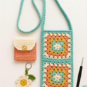 Op de afbeelding: Een verzameling handgemaakte haakartikelen, waaronder een turquoise crossbodytas, een klein perzikkleurig etui en een sleutelhanger met madeliefjes. De tas en het etui hebben kleurrijke vierkante patronen. Een haaknaald en garen zijn ook aanwezig.