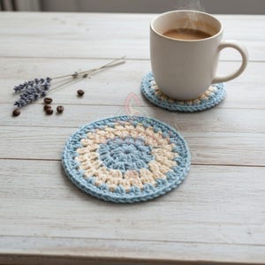 May include: A handmade crochet coaster with a blue, cream, and light pink spiral design. A white mug filled with coffee sits on a matching coaster. Lavender sprigs and coffee beans are also on the light wood surface.