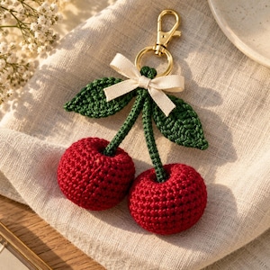 May include: A crochet cherry keychain featuring two round, textured red yarn cherries attached to green stems and leaves. A small, off-white fabric ribbon is tied at the top, connecting to a gold-toned metal lobster clasp for easy attachment.