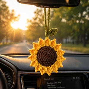May include: A crocheted sunflower car charm with yellow petals and a brown center, hanging from a green stem and leaf. The charm is suspended from a car's rearview mirror, with a dashboard and a sunlit road in the background.