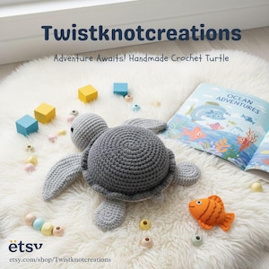 Puede incluir: Una tortuga de ganchillo gris, con caparazón y aletas texturizadas, descansa sobre una superficie blanca y esponjosa. La imagen incluye un libro infantil titulado "Ocean Adventures", bloques de madera de colores y el texto "Twistknotcreations".