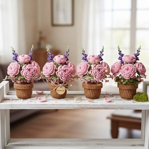 May include: Four crochet flower arrangements in brown pots sit on a white shelf. Each arrangement features pink roses and purple flowers. A small wooden tag with the word "handmade" is tied to one pot.