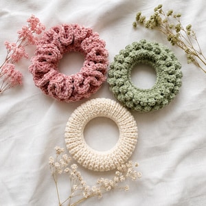 May include: Three handmade scrunchies in shades of pink, green, and cream, arranged on a white surface. The pink scrunchie has a velvet texture, the green one has a textured, bobbled design, and the cream one is knitted. Dried flowers are scattered around the scrunchies.