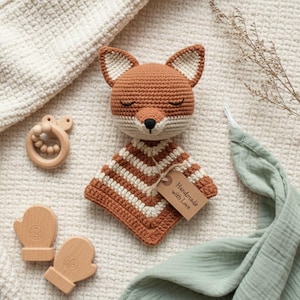 Könnte beinhalten: Gehäkelter Fuchs-Baby-Kuscheltier mit braun-weiß gestreifter Decke. Der Fuchs hat geschlossene Augen und eine schwarze Nase. Ein Holzbeißring und zwei Holzfäustlinge sind ebenfalls im Bild. Ein Etikett lautet "Handmade with Love."