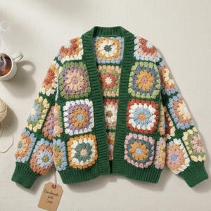May include: A colorful, open-front crochet cardigan. The sweater features a patchwork design with square motifs in various pastel colors, framed by a dark green border and trim. The tag reads "Handmade with Love".