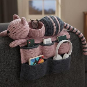 May include: A pink and gray crocheted cat-shaped organizer with multiple pockets. The organizer has a cat head, a striped tail, and pockets for items like glasses, a remote, and headphones. The design is soft and whimsical.