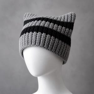 May include: A crochet beanie hat featuring a cat-ear silhouette with a ribbed texture. The design consists of alternating horizontal stripes in solid grey and black yarn, creating a distinct pattern across the entire surface of the headwear.