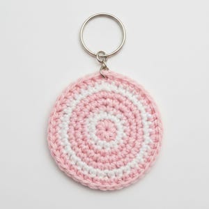 May include: A handmade, circular keychain featuring a crocheted design in pink and white concentric circles. The keychain has a silver metal ring for attaching keys. The crochet work is detailed and the color scheme is soft and appealing.