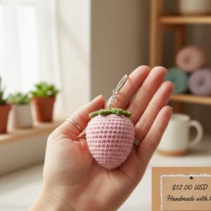 May include: A crocheted strawberry keychain in light pink with a green leafy top. The handmade accessory is held in a hand, with a silver keyring attached. The background includes small potted plants and a window.