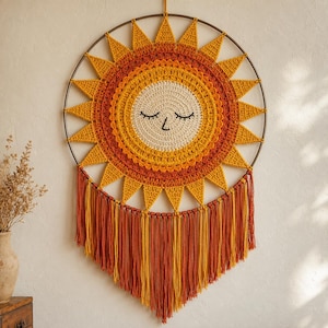 May include: A circular wall hanging featuring a crocheted sun design with a cream-coloured face, closed eyes, and a stitched nose. The sun has radiating yellow and orange triangular rays attached to a metal hoop, with long orange and yellow fringe.