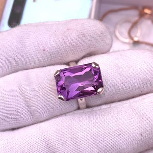 Op de afbeelding: Een zilveren ring met een grote, rechthoekige, gefacetteerde paarse edelsteen. De edelsteen is gezet in een zilveren band met kleine zilveren accenten op elke hoek. De ring wordt gepresenteerd op een witte stoffen ondergrond.