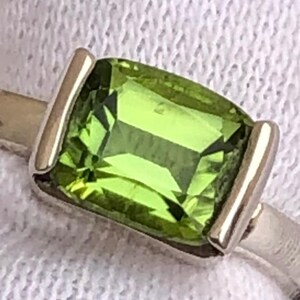 May include: A silver ring with a rectangular, light green gemstone. The gemstone is set between two silver bars, creating a modern design. The ring is displayed on a white, textured surface.
