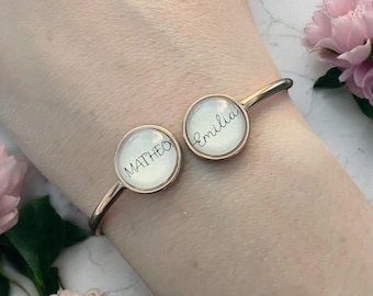 Op de afbeelding: Een zilverkleurige open manchetarmband met twee cirkelvormige bedels. Elke bedel heeft een witte achtergrond met een naam in zwart schrift: "MATHEO" en "Emilia". De armband wordt getoond op een pols met roze bloemen op de achtergrond.