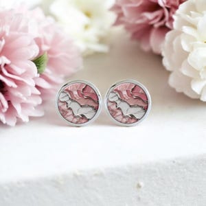 Pode incluir: Um par de abotoaduras redondas prateadas com um design abstrato rosa, branco e cinza. As abotoaduras são apresentadas sobre um fundo de flores rosa e brancas, criando uma estética delicada e elegante.
